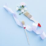 Two paint brushes with ribbon and flowers on a pastel blue background, ideal for DIY and decor themes.