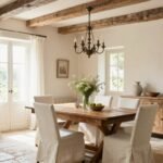 A bright, airy French country dining room with a large wooden farmhouse table, linen-upholstered chairs, and a vintage chandelier hanging above