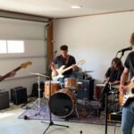 A garage band rehearsing with guitars, drums, and microphones in a garage setting